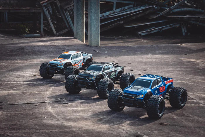 Three remote-controlled FMS 1/24 Chevrolet Colorado monster trucks on a concrete surface with industrial background