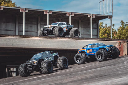 Three FMS 1/24 Chevrolet Colorado RC with LED lights, monster trucks on a track with a building and trees in the background