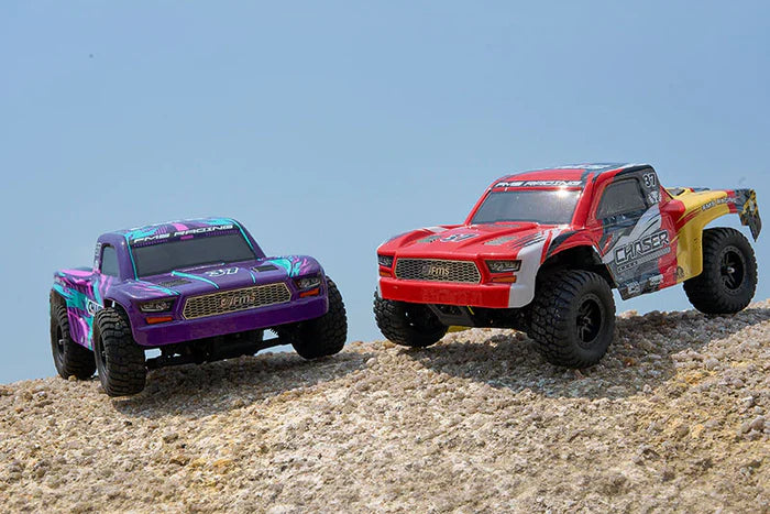 Two Chaser 1/24 RC off-road trucks, one purple and one red with yellow accents, on a sandy surface.
