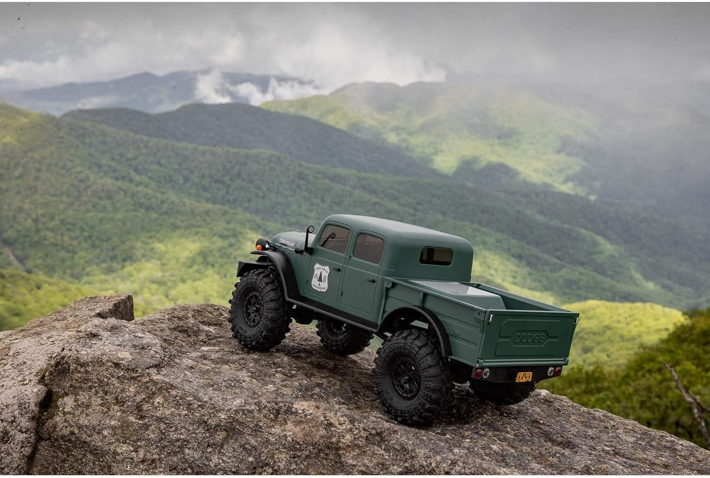 Green off-road Axial 1/24 SCX24 Dodge RC mini off-road crawler on a rocky outcrop with misty mountains in the background