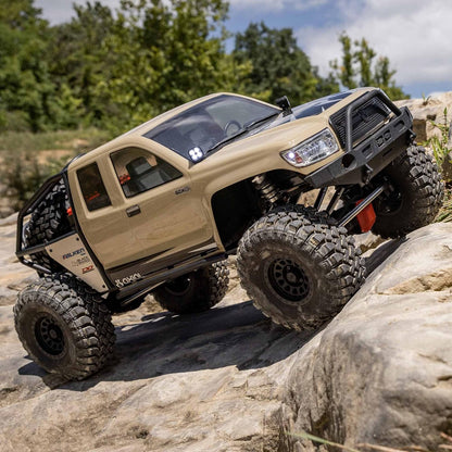 Axial SCX6 Trail Honcho 1/6 scale 4WD RTR rock crawler – beige off-road vehicle climbing a rocky terrain with trees in the background, massive tires, roll cage, high-clearance chassis