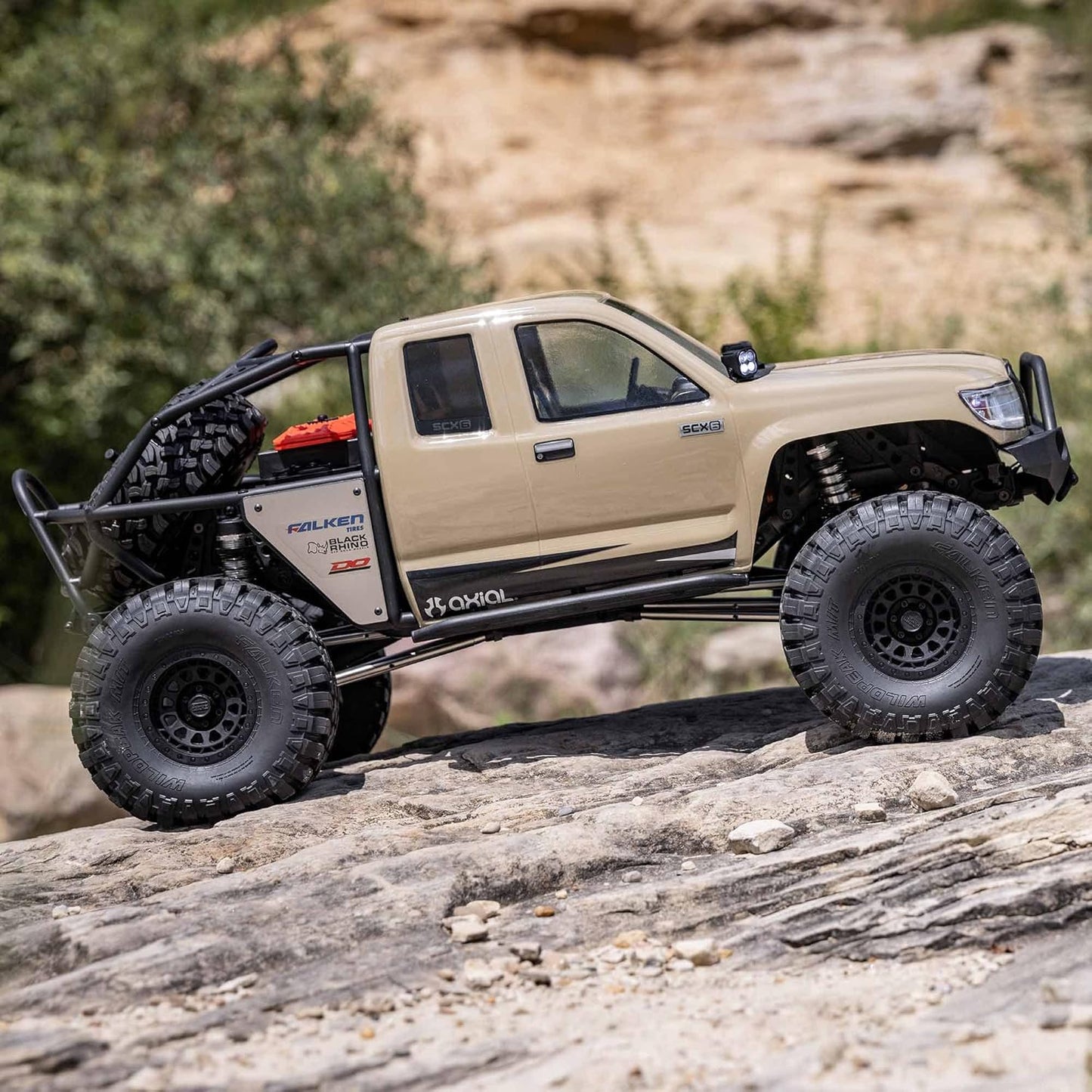 1/6 scale Axial SCX6 Trail Honcho RC crawler – beige off-road vehicle climbing rocky terrain with trees background, detailed suspension, realistic scale