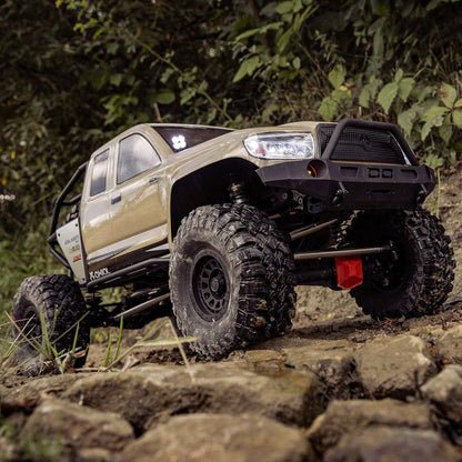 Axial SCX6 Trail Honcho 1/6 scale 4WD RTR rock crawler, off-road vehicle on a rocky terrain with greenery in the background, beige body, massive tires, roll cage, and high-clearance chassis